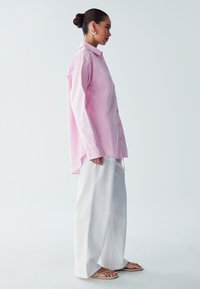 Camicia button-up rosa chiaro con maniche lunghe, abbinata a pantaloni bianchi larghi e sandali piatti. Tessuto liscio, vestibilità rilassata e colletto classico.