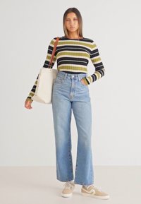 Striped strikket topp i grønn og svart, høy midje lys blå jeans, beige sneakers, og en kremfarget tote bag, står foran en ensfarget bakgrunn.