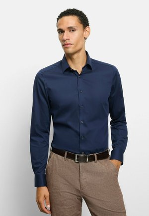 Marineblaues Hemd mit Knöpfen und langen Ärmeln, gefertigt aus glattem Stoff. Kombiniert mit hellbraunen, strukturierten Hosen und einem dunklen Gürtel.