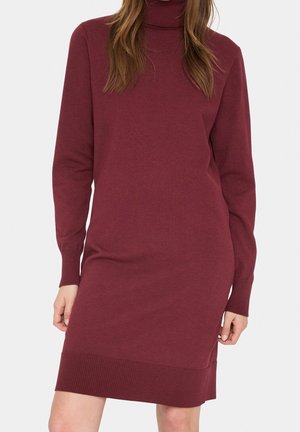 Femme portant une robe tricotée bordeaux à manches longues avec un ourlet côtelé et un col montant, se tenant devant un fond blanc uni.