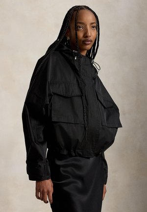 Femme aux longs cheveux tressés portant une veste noire ample avec de grandes poches avant et une jupe noire sur un fond uni.