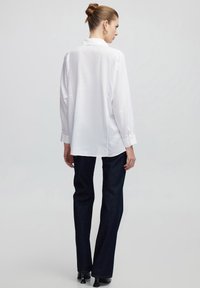 Camicia bianca con bottoni, dalla vestibilità rilassata, con maniche lunghe e una piega posteriore, abbinata a pantaloni flair in denim scuro e tacchi neri.