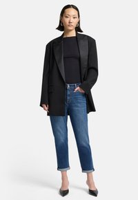 Blazer nero con rever in satin su una canotta nera, abbinato a jeans in denim blu arrotolati e tacchi a punta neri.