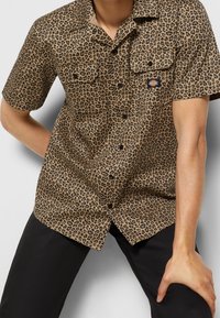 Koszula z krótkim rękawem wykonana z brązowego materiału w czarno-leopardzi wzór, z dwoma kieszeniami na piersi i zapięciem na guziki.