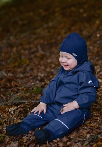 Combinezon impermeabil bleumarin cu glugă, cu manșete elastice și accente reflectorizante albe, purtat de un copil așezat pe frunze uscate.