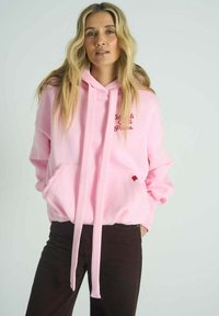 Růžová mikina s kapucí z fleecového materiálu s přední kapsou. Má dlouhé šňůrky a červený textový motiv. Kombinována s tmavě hnědými kalhotami.