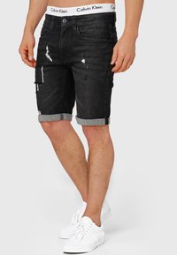 Shorts di denim neri con orli risvoltati, caratterizzati da toppe consumate e un elastico in vita con il marchio Calvin Klein. Indossati con sneakers bianche.