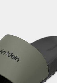 Tuvējs skats uz olīvu zaļās un melnās slaidu sandalēm ar iespiestu Calvin Klein logotipu uz siksnas.