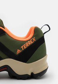 Scarpa sportiva verde con accenti arancioni, tomaia in rete, striscia nera e suola testurizzata. Logo "Terrex" visibile sul lato.