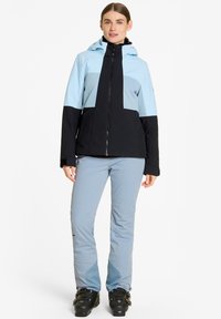 Blaue und schwarze Skijacke mit Kapuze, die ein Farbblockdesign aufweist. Kombiniert mit hellblauen Skihosen und schwarzen Skischuhen.
