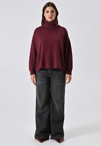 Burgunderfarbener Rollkragenpullover mit lockerer Passform, kombiniert mit hochgeschnittenen schwarzen Weitbeinjeans. Glatte Textur mit geripptem Kragen.