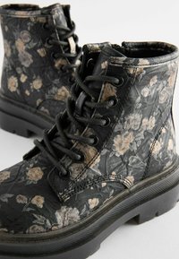 Bottines noires à motif floral, avec une texture mate, un bout rond, une semelle épaisse, et des lacets noirs avec des œillets métalliques.
