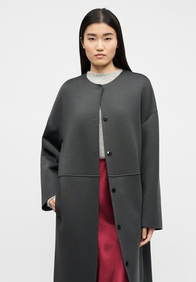 Μια γυναίκα με μακριά σκουρόχρωμα μαλλιά φοράει ένα μακρύ σκούρο γκρι παλτό με κουμπιά πάνω από ένα ανοιχτό γκρι τοπ και κόκκινη φούστα, στέκεται μπροστά σε ένα απλό φόντο.