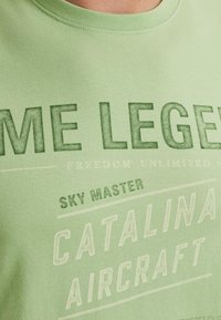 T-shirt en coton vert clair avec un texte audacieux en vert foncé et blanc, comprenant "LÉGENDE" et "CATALINA AIRCRAFT", avec un col rond classique.