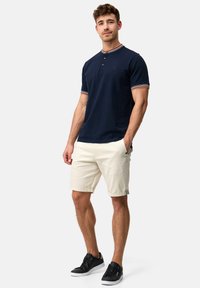 Marine polo skjorte med kontrasterende stribet krave, tre knapper, bæres med cremede shorts og sorte sneakers. Stof ser glat og afslappet ud.