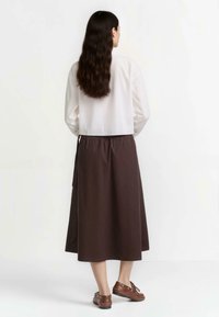 Bluză albă cu mâneci lungi, purtată cu o fustă închisă la culoare, până la gleznă, cu talie elastică și accente legate. Pantofi maro completează look-ul.