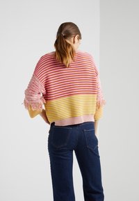 Frau mit braunen Haaren im Pferdeschwanz, die einen gestreiften Pullover in Pink, Gelb und Weiß mit Fransen an den Ärmel und eine dunkle Jeans trägt, mit dem Rücken zur Kamera.