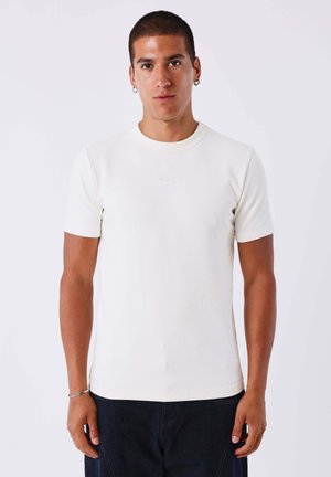 Persoon met kort donker haar die een effen wit T-shirt met korte mouwen en een donkere broek draagt, staat met het gezicht naar voren tegen een lichte achtergrond.