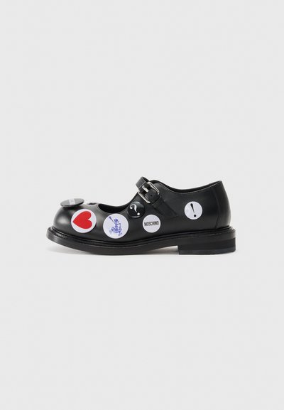 Chaussure Mary Jane en cuir noir avec une sangle et une boucle, ornée de divers autocollants ronds aux accents rouges, bleus et blancs. Design à semelle plate.