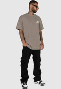 Tricou oversized din bumbac gri, cu un grafic mic al unei muntii albi, asortat cu pantaloni cargo negri cu buzunare și adidași negri și albi.