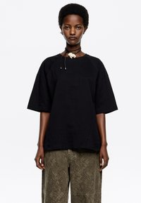 T-shirt nera oversize con maniche raglan, realizzata in tessuto morbido. Abbinata a pantaloni verde oliva a fantasia.