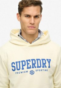 Felpa color crema con un grande logo blu "SUPERDRY". Presenta un cappuccio con cordino e una texture morbida. Indossata sopra una camicia con colletto.