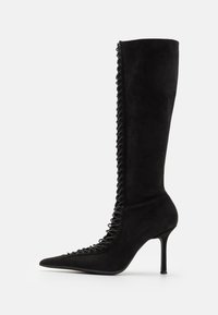Boot cu toc înalt, din suede negru, de lungime până la genunchi, cu vârf ascuțit, având detalii din dantelă pe lateral și o siluetă elegantă.