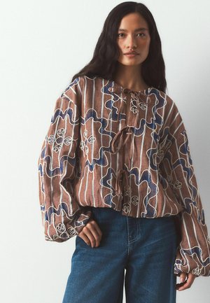 Žena nosi voľnú hnedú blúzku s modrým a bielym kvetinovým vyšívaním a vertikálnymi pruhmi, doplnenú o vysoký pás modrých džínsov.