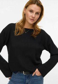 Melns garroku tops no mīksta materiāla, ar vaļīgu piegriezumu un platiem aprocēm, kombinēts ar zilām džinsa biksēm un melnu jostu.