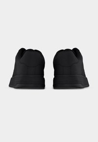 Ζευγάρι μαύρα αθλητικά παπούτσια χαμηλού ύψους, εμφανιζόμενα από πίσω, με λεία υφή και λαστιχένιες σόλες σε απλό φόντο.