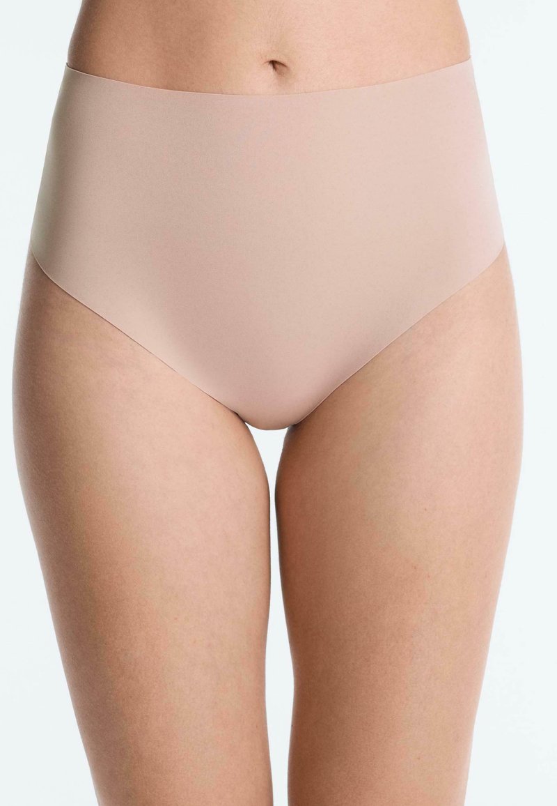 Femme portant des sous-vêtements nude sans coutures taille haute, montrant le torse avant et le haut des cuisses sur un fond uni.