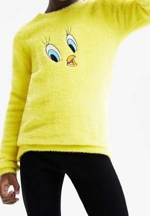 Person iført en blød gul sweater med et stort broderet Tweety Bird-ansigt på fronten, kombineret med sorte bukser.