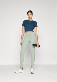 Tengerész kék, rövidített póló diszkrét logóval, világoszöld, magas derekú leggings és fehér sportcipő; egy személy fekete vizespalackot tart.