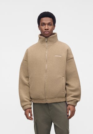 Un homme porte une veste polaire beige à col montant, avec une fermeture éclair sur le devant et des poches, associée à un pantalon cargo foncé sur un fond uni.