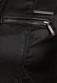 Nærbillede af sort stof med en lille vandret lynlås og et metalmærke fra "Guess" over lynlåsen.