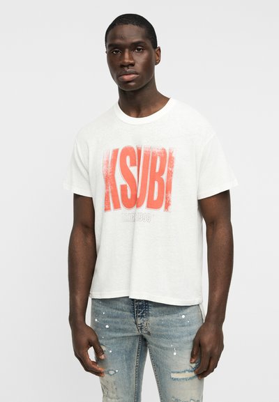 Λευκό T-shirt με κοντά μανίκια, με έντονο κόκκινο λογότυπο "KSUBI" με εφέ φθοράς, σε συνδυασμό με ανοιχτόχρωμο μπλε τζιν με φθορές.