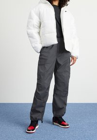 Vit puffjacka med dragkedja, kombinerad med mörkgrå cargobyxor med sidofickor, och svart-röda sneakers med vita sulor.