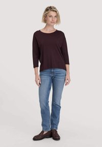 Weinrote Langarmbluse mit rundem Ausschnitt, kombiniert mit hellblauen Jeans; das Model trägt braune geflochtene Slipper.