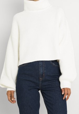 Personne portant un pull à col roulé blanc côtelé avec des manches bouffantes et un jean taille haute bleu foncé sur un fond uni.
