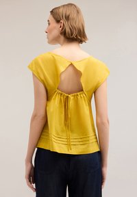 Blusa gialla a maniche corte con un'apertura sulla schiena e dettaglio con fiocco arricciato. Pannelli texturizzati vicino all'orlo. Abbinata a denim scuro.