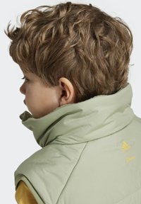 Gilet imbottito verde chiaro con colletto alto, con logo giallo Adidas e Disney. Texture morbida con una finitura leggermente lucida.