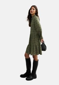 Robe vert olive à pois noirs, design à volants, manches longues et col, associée à des bottes noires jusqu'aux genoux et à un petit sac.