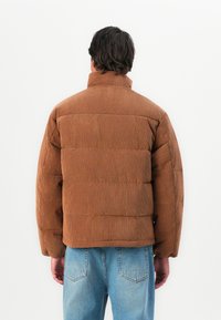 Man gekleed in een bruin corduroy pufferjack en blauwe jeans, staande met zijn rug naar de camera tegen een effen witte achtergrond.