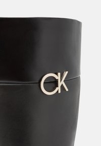 Stivale in pelle nera con una texture liscia, caratterizzato da un prominente logo "CK" in tono oro sul lato e un design elegante e aderente.