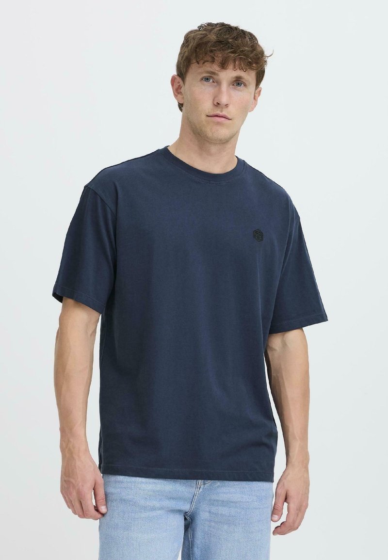 Camiseta de algodón azul marino con cuello redondo, mangas cortas y un pequeño logo hexagonal negro en el lado izquierdo del pecho. Textura suave.