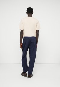 Tricou bej cu mânecă scurtă și pantaloni marin cu un buzunar la spate. Modelul stă cu spatele la o fundal alb, purtând pantofi maron.