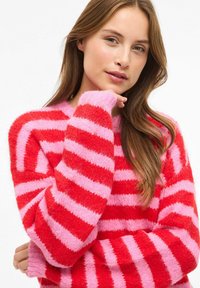 Młoda kobieta z długimi brązowymi włosami, ubrana w czerwoną i różową sweterkową bluzę w paski, opierająca podbródek na dłoni na białym tle.