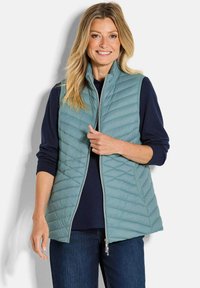 Lächelnde Frau in einer hellblauen Steppweste über einem marineblauen Langarmshirt und blauen Jeans, stehend vor einem schlichten Hintergrund.