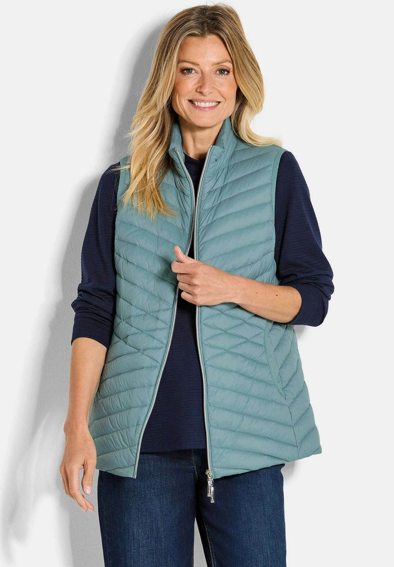 Lächelnde Frau in einer hellblauen Steppweste über einem marineblauen Langarmshirt und blauen Jeans, stehend vor einem schlichten Hintergrund.