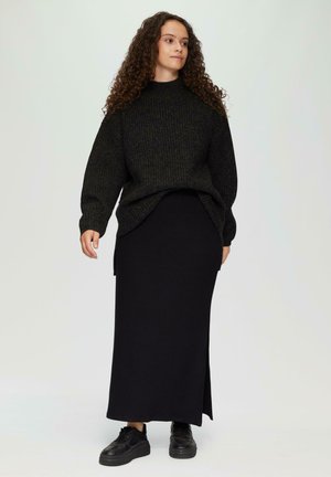 Schwarzer gestrickter Pullover mit hohem Kragen, lockerer Passform und gerippter Textur, über einem langen schwarzen Rock mit seitlichem Schlitz getragen, kombiniert mit schwarzen Sneakers.
